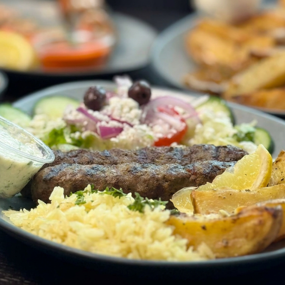 Grilled kofta served with rice, roasted potato wedges, salad, lemon, and creamy herb sauce on a dark plate.