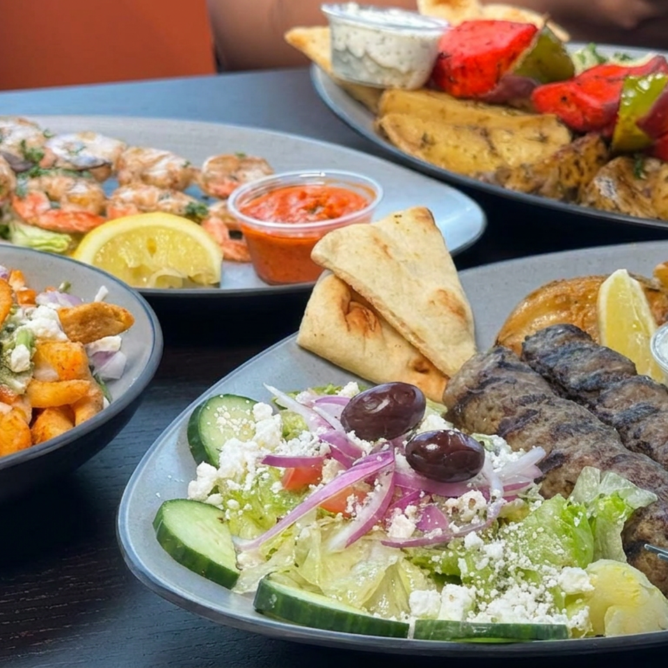 Greek-style feast featuring grilled kebabs, shrimp skewers, pita, Greek salad, roasted vegetables, rice, sauces, and lemon wedges.